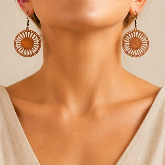 Close-up of a woman wearing large circular copper-toned earrings with radiating cutout details and dark hooks against a warm beige background.