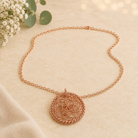 A rose gold necklace with a circular, intricate floral wire pendant, displayed on a textured beige fabric background with soft bokeh lights and eucalyptus flowers in the corner.