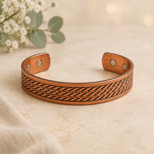 A copper cuff bracelet with an engraved basketweave pattern, displayed on a light beige textured surface with soft bokeh background lights and small white flowers in the corner.
