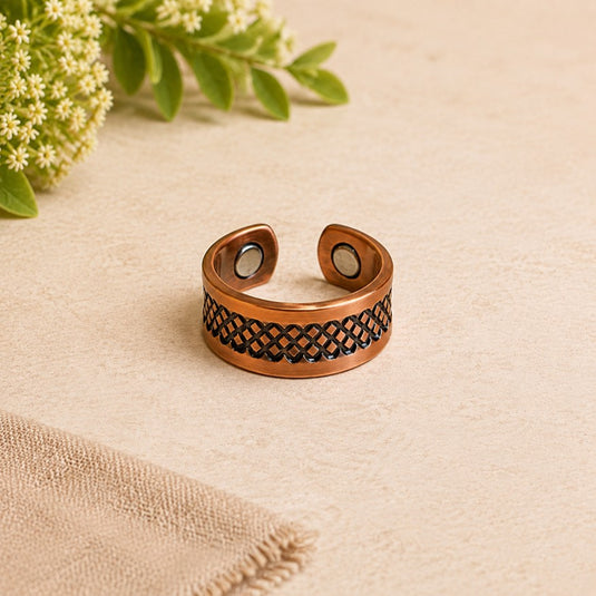A copper-toned open cuff ring with a black woven pattern band and circular magnetic ends, styled on a beige textured surface with small white flowers and green leaves in the corner.