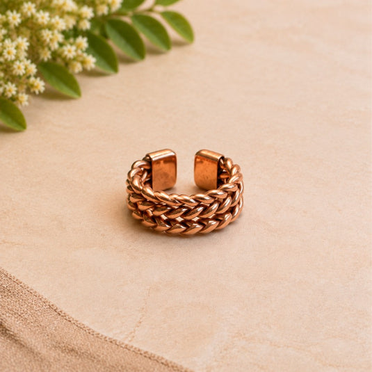 A copper-toned open cuff ring with a braided textured band, displayed on a beige textured surface with small white flowers in the upper left corner.
