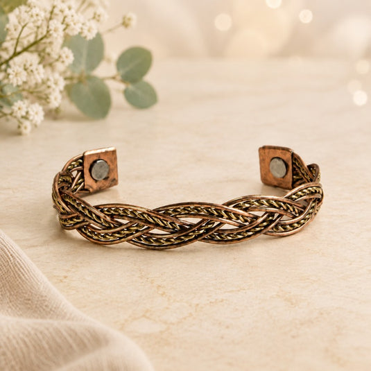 A braided metallic cuff bracelet with a warm copper-bronze finish and square magnetic ends, styled on a light beige textured surface with soft floral props in the background.
