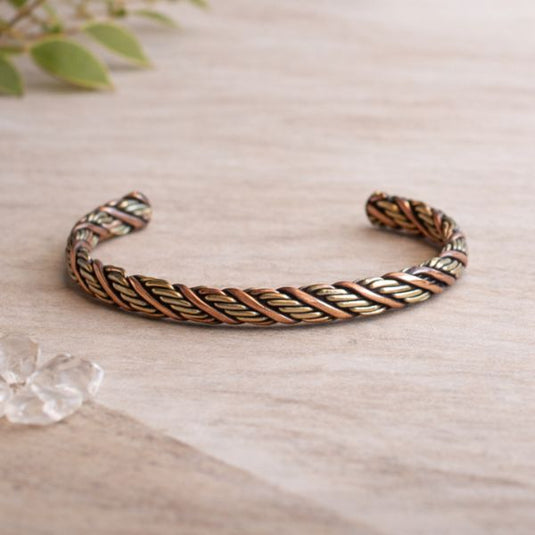 Woven metal bracelet on a light wooden surface with greenery in the background