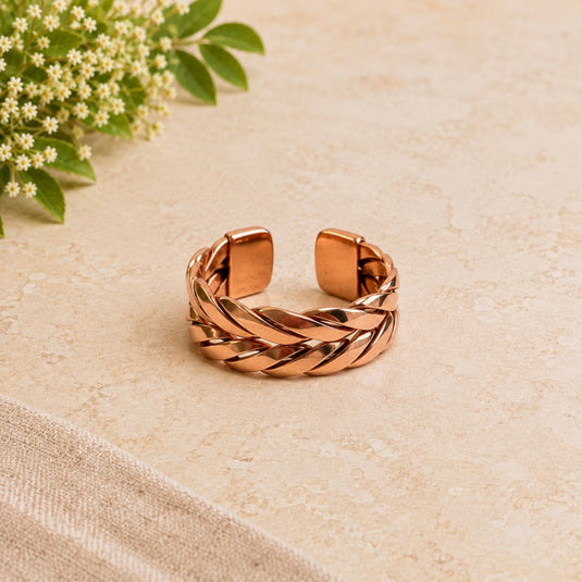 A copper-toned twisted braid cuff bracelet with a polished finish, displayed on a textured beige surface with a small green floral sprig in the corner.
