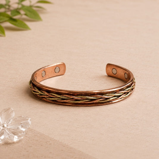 Copper and gold braided bracelet on a beige surface with green leaves in the background