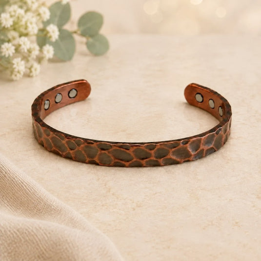 A hammered copper-toned metal cuff bracelet with three silver-tone rivet details on each end, displayed on a textured beige surface with soft-focus eucalyptus and white flowers in the background.